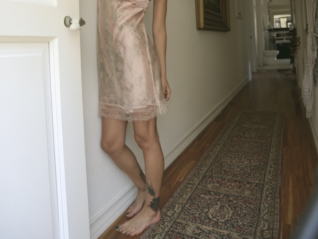 Barefoot woman in pink satin slip with leg tattoo standing in narrow hallway on vintage runner rug