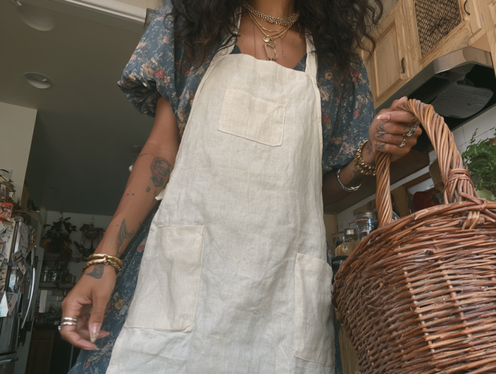 Tattooed home cook in floral dress and linen apron holding wicker basket in rustic kitchen