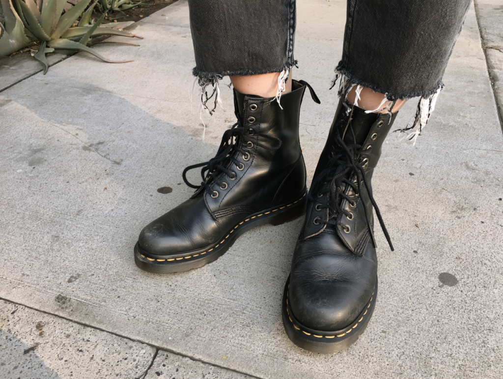 Black lace-up combat boots with yellow stitching and chunky soles, paired with distressed hem jeans on a sidewalk.