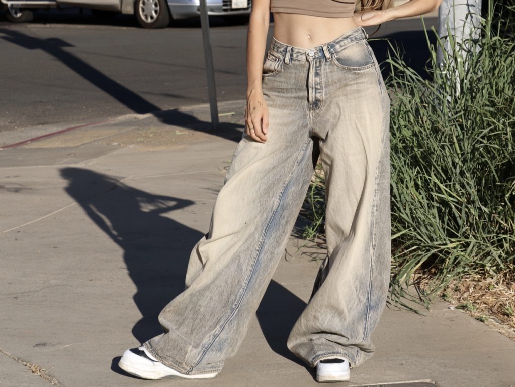 Model in beige crop top wearing baggy wide-leg light-wash jeans and white sneakers on city sidewalk, street style.