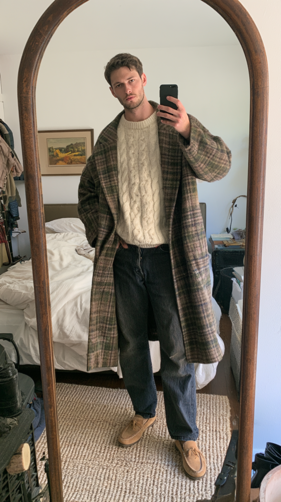 winter-old-money-outfits-inherited-wool-coat Man in bedroom mirror selfie wearing plaid overcoat, cream cable-knit sweater, dark jeans, and moccasins; cozy fall outfit.