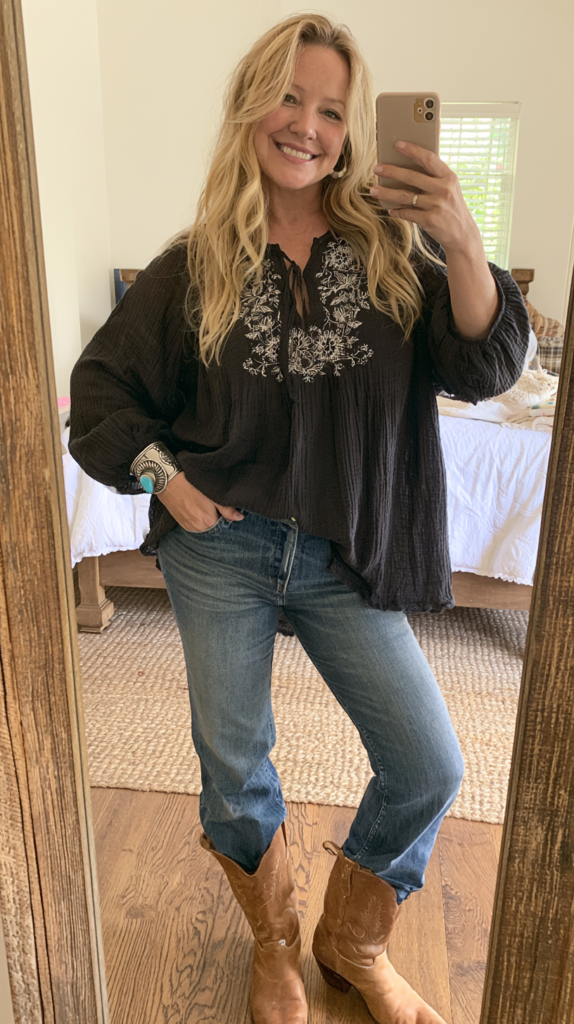 Mirror selfie of woman in black embroidered boho blouse, blue jeans, and brown cowboy boots, western casual outfit idea.